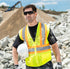 Construction worker in safety vest and sunglasses holding a hard hat in front of a construction site.